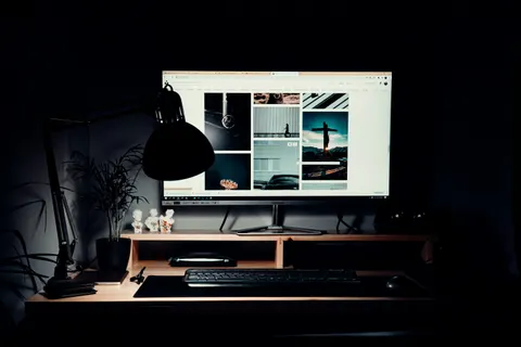 Poste de travail d'un développeur web la nuit : grand écran allumé sur une galerie de sites, lampe d'architecte, clavier et plante posés sur un bureau en bois