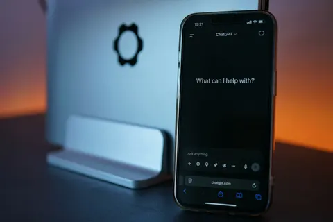 Smartphone ouvert sur ChatGPT affichant « What can I help with? », posé devant un ordinateur, le point de départ de qui veut créer son site avec l'IA