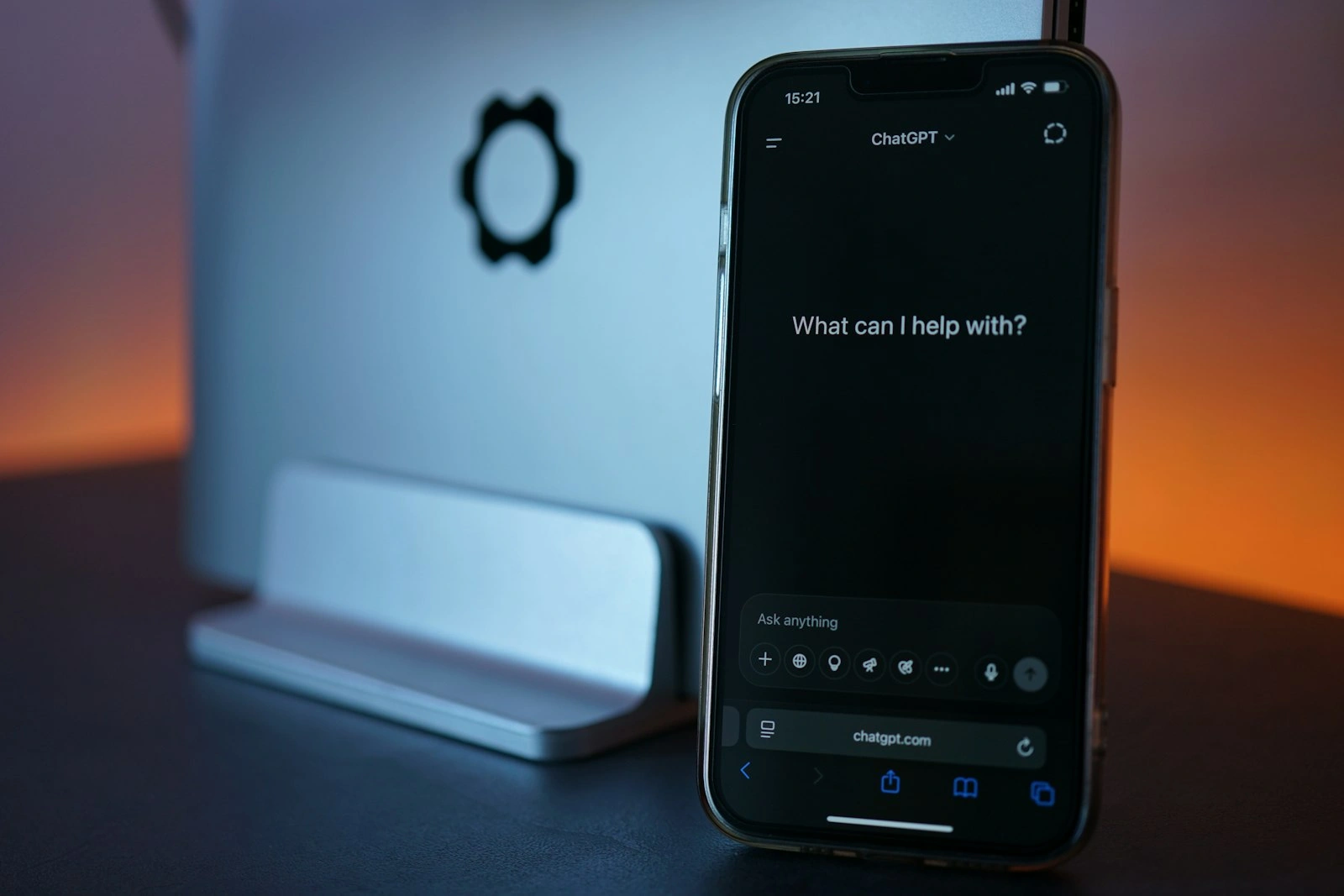 Smartphone ouvert sur ChatGPT affichant « What can I help with? », posé devant un ordinateur, le point de départ de qui veut créer son site avec l'IA