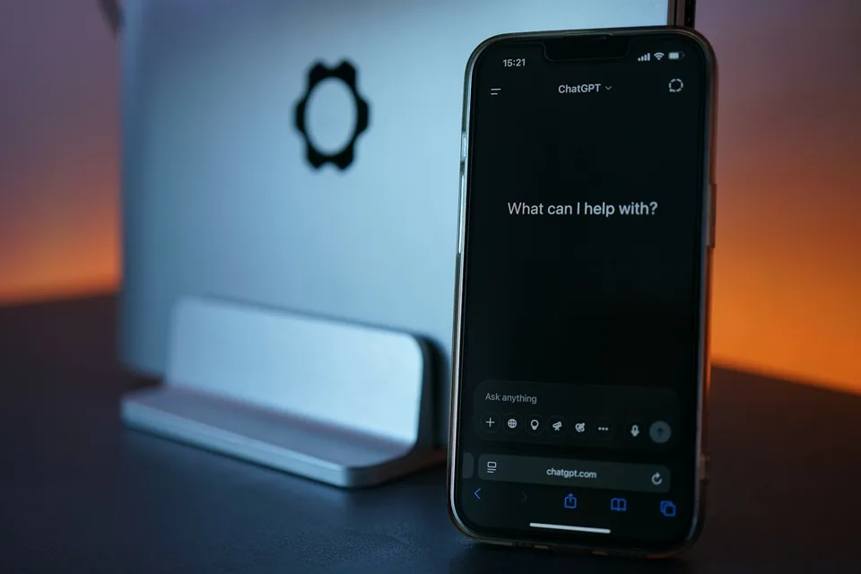 Smartphone ouvert sur ChatGPT affichant « What can I help with? », posé devant un ordinateur, le point de départ de qui veut créer son site avec l'IA