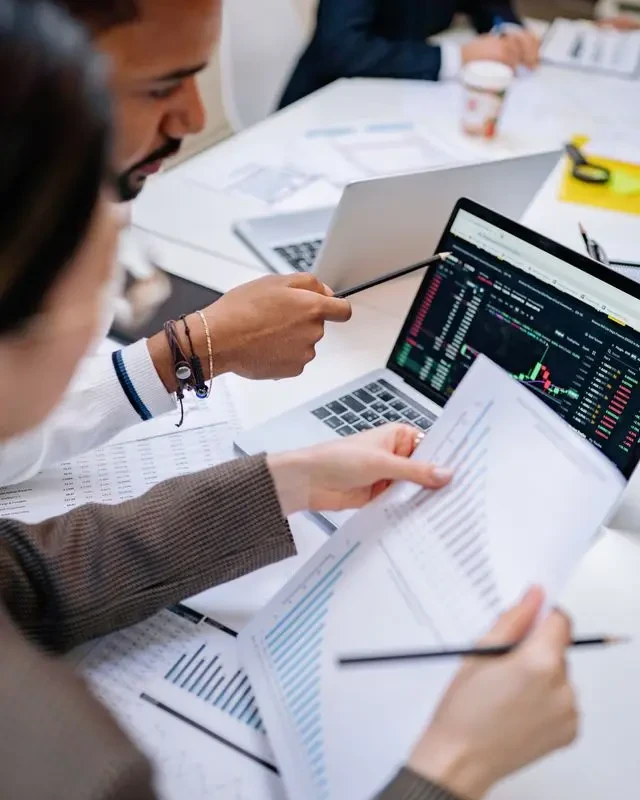 Deux personnes analysant des graphiques financiers imprimés à côté d’un graphique boursier sur ordinateur